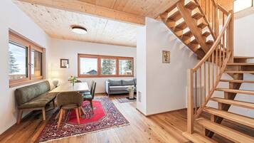 A spacious living area with a wooden staircase, a sofa, a dining table with chairs, and large windows.