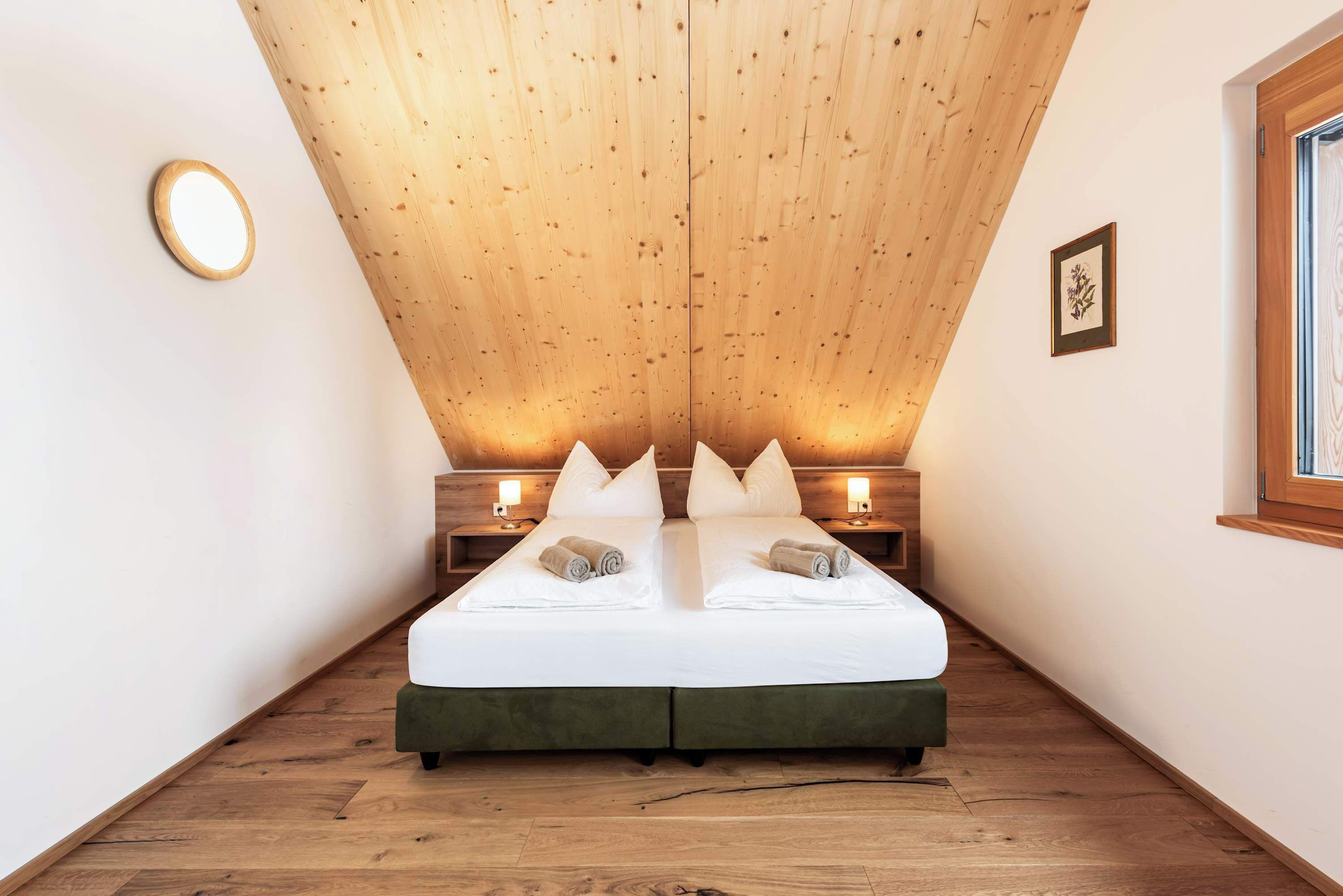 A bedroom with a wooden sloped ceiling, a bed with white linens, two bedside tables with lamps, and a framed picture on the wall.