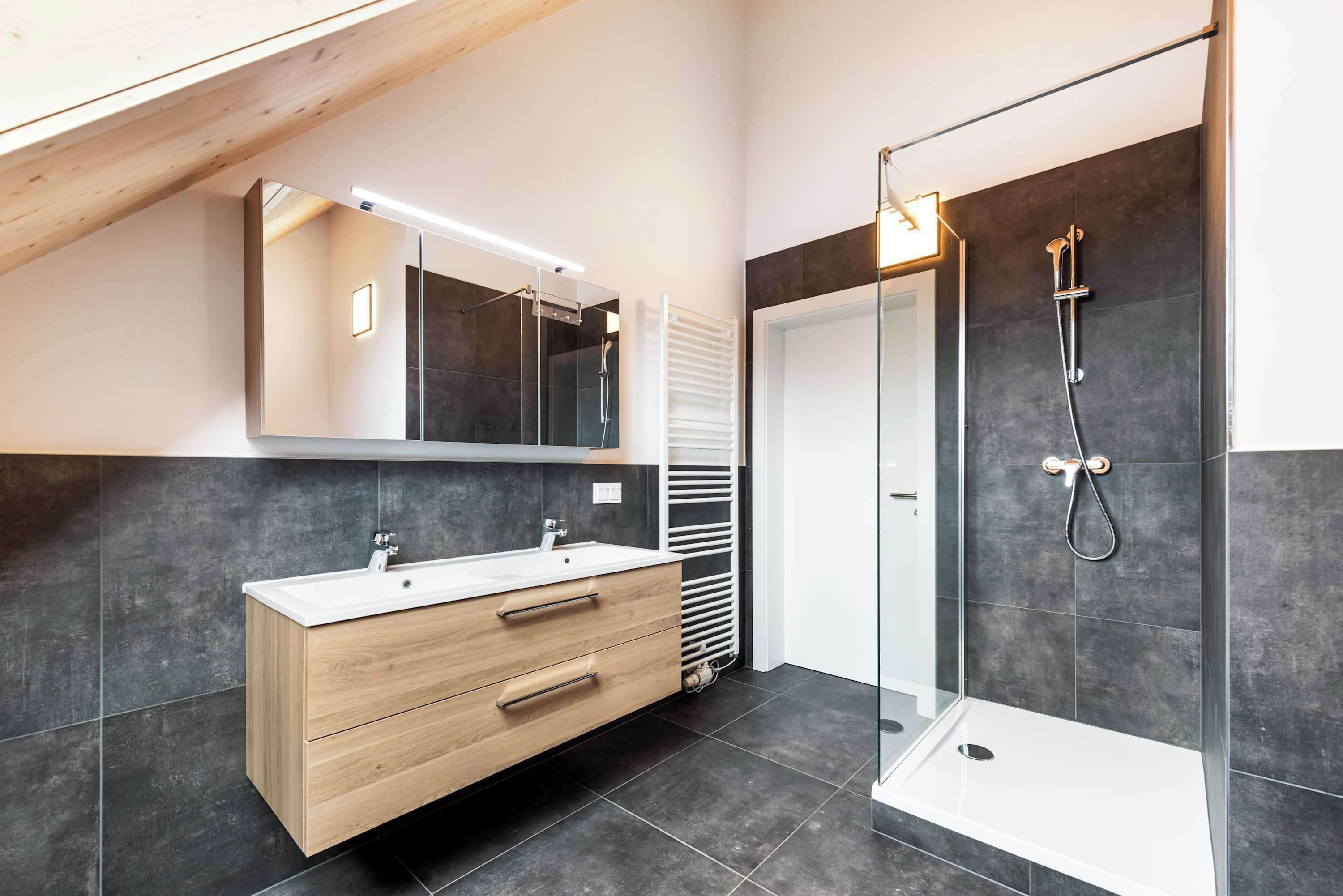 A modern bathroom with a wooden vanity, a large mirror, a glass shower enclosure, and dark tiled walls.