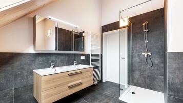 A modern bathroom with a wooden vanity, a large mirror, a glass shower enclosure, and dark tiled walls.