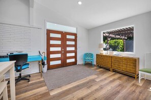 A modern room with a wooden floor, a desk with a computer, a blue chair, a cabinet, and a window with a view of greenery. - Heated Pool • Pet Friendly • Guest House • Walk to Village (St simons)