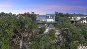 Une maison à deux étages dotée d’un balcon, entourée d’une végétation luxuriante et d’un terrain rocheux.