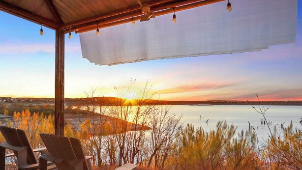 Un espace extérieur aménagé, abrité et doté de chaises, offrant une vue sur un coucher de soleil se reflétant sur une étendue d’eau.