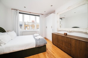 A modern bedroom with a large bed, a wooden dresser, a mirror, and a window with curtains. - Flat 3 Elegant 2 Bed Marylebone Flat with Balcony (London)