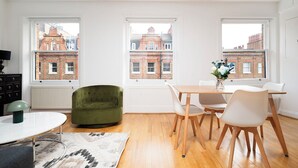 Een moderne woonkamer met een groene fauteuil, een eettafel met stoelen en grote ramen met uitzicht op gebouwen van rode baksteen.