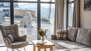 Un salon moderne avec un canapé, un fauteuil et une table basse en bois. De grandes fenêtres offrent une vue sur les montagnes et un jardin de sculptures en pierre.