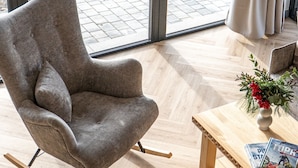 Une pièce moderne avec un fauteuil à bascule à revêtement gris, une table basse en bois sur laquelle sont posés des magazines, et une vue sur un espace extérieur grâce à de grandes fenêtres.