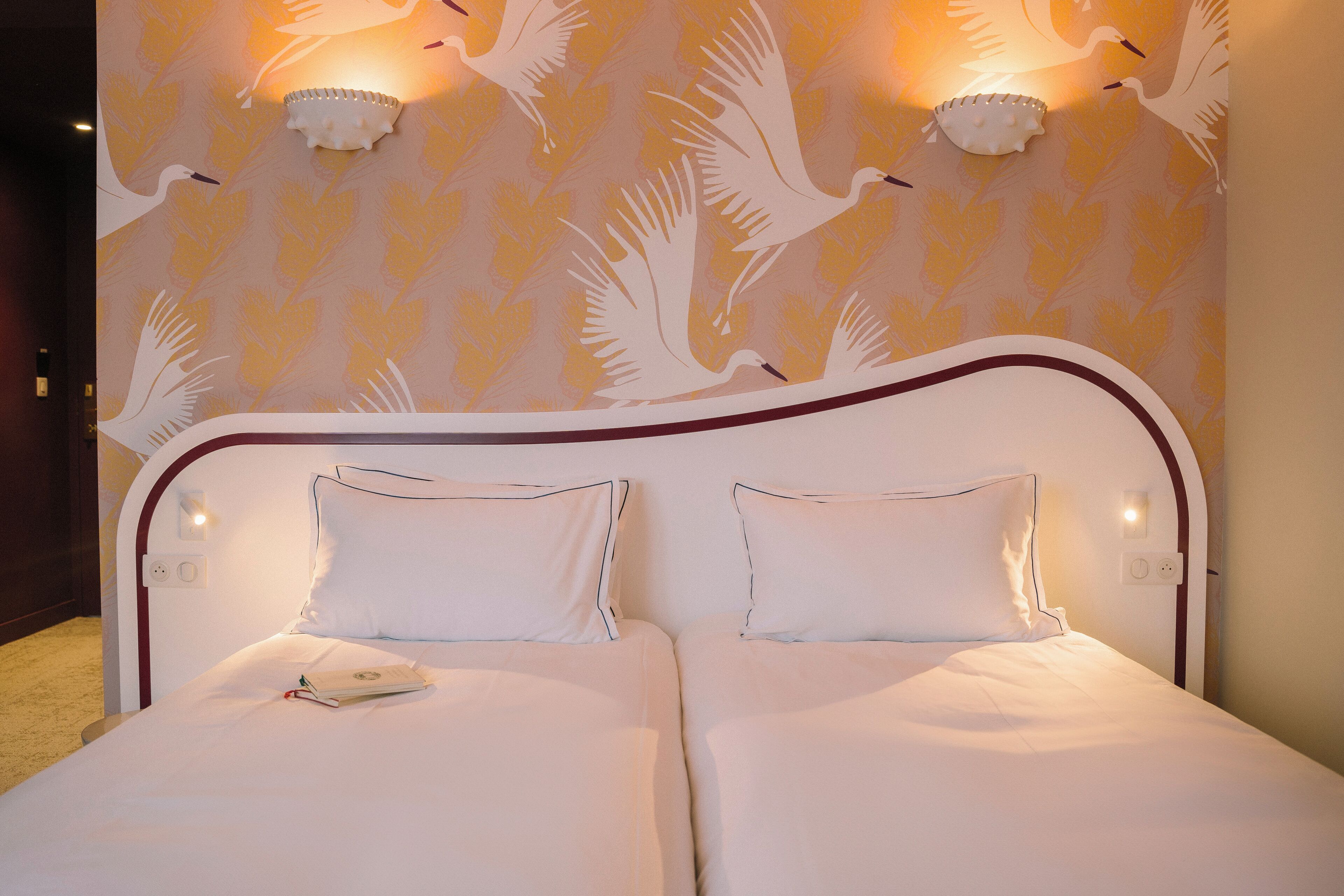 Una cama doble con ropa de cama blanca y dos almohadas, una mesita de noche y apliques de pared con un motivo de pájaros.