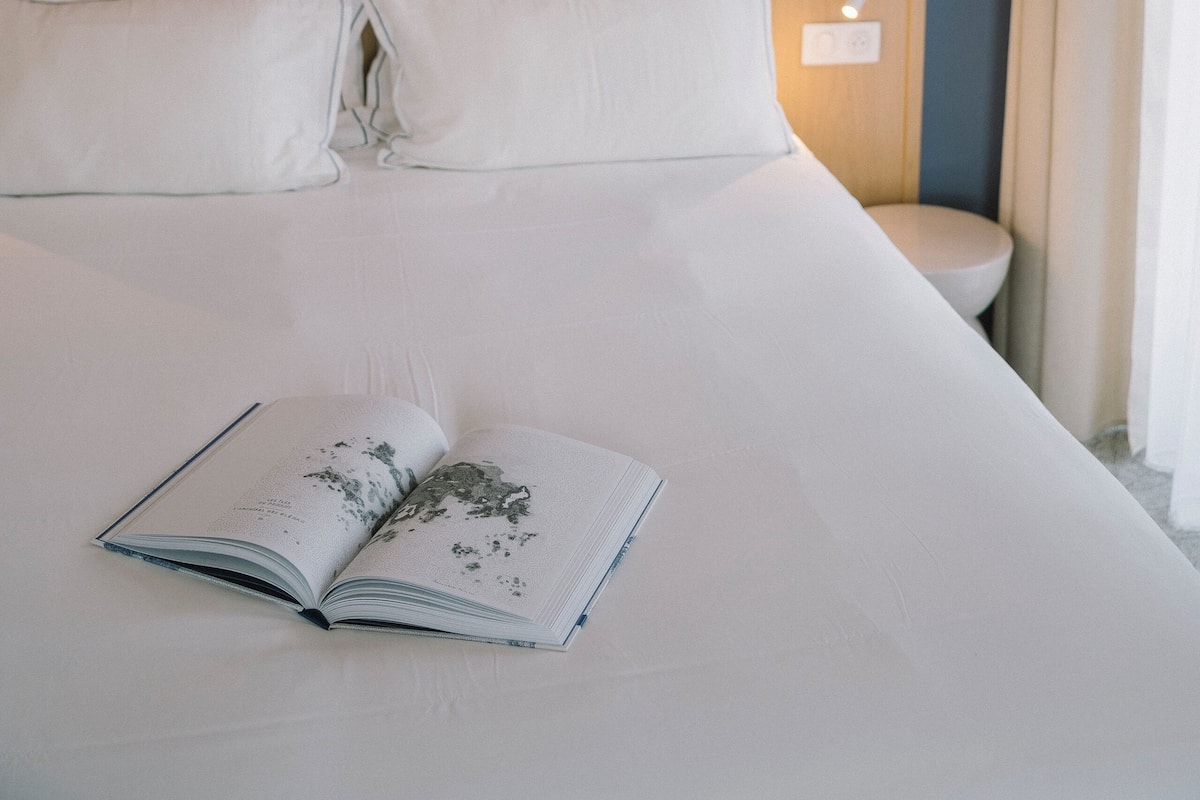 Una cama bien hecha con un libro encima, un cuadro en la pared y una mesita de noche con una lámpara.