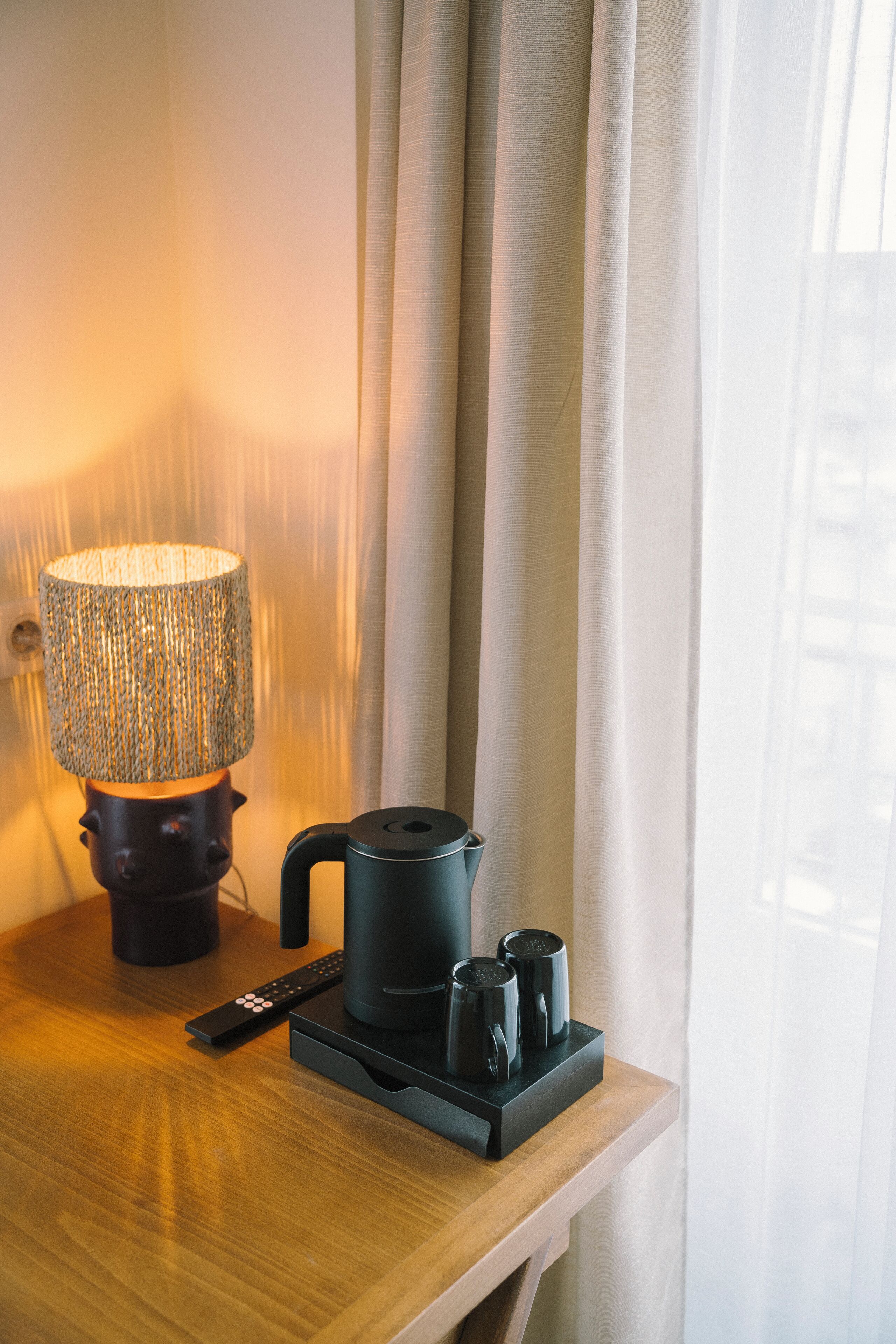 A wooden side table with a lamp, remote control, and a coffee maker with cups.