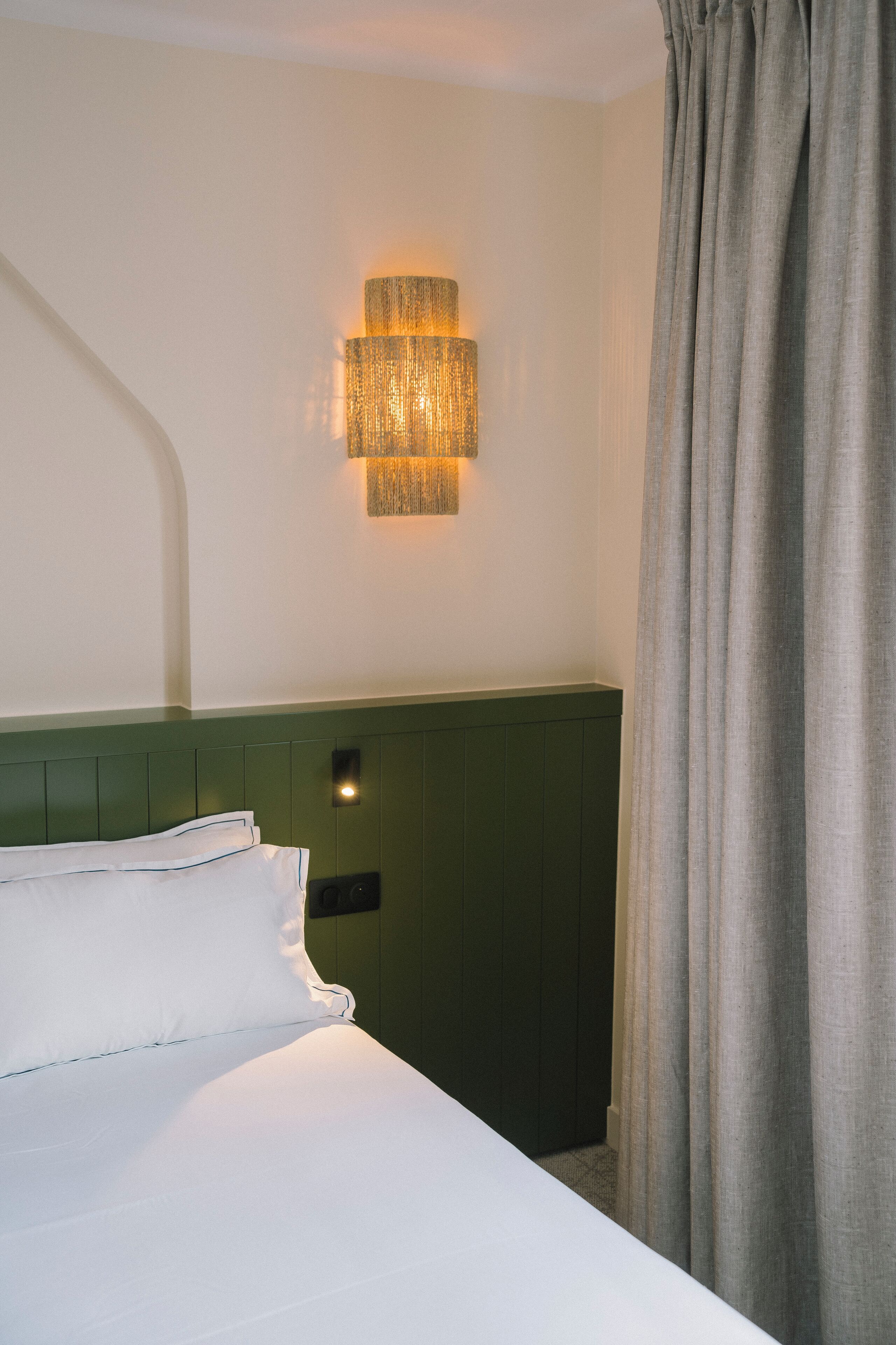 Una habitación de hotel con cama, lámpara de pared y cabecero verde.