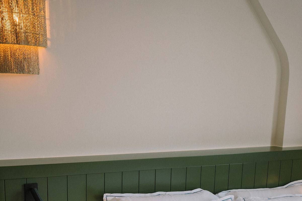 Una cama bien hecha con sábanas y almohadas blancas, un cabecero verde y una lámpara fijada a la pared.