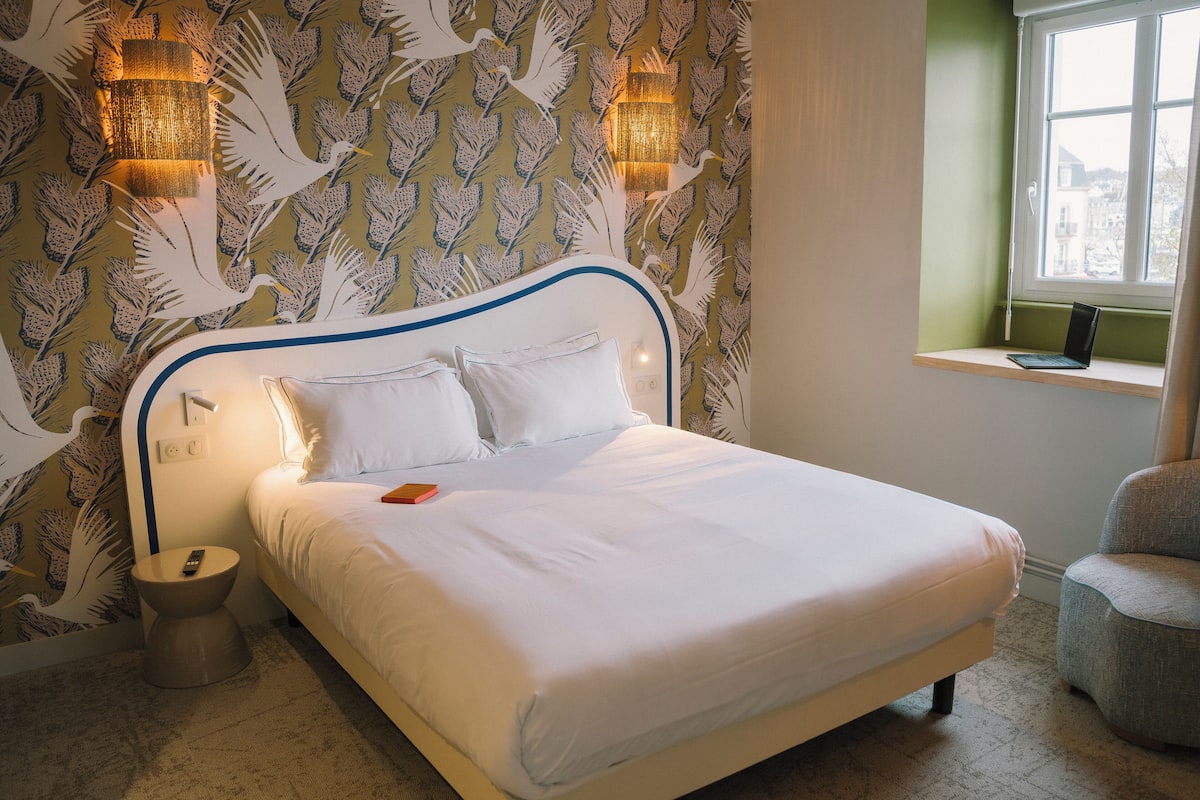 Una cama bien hecha con sábanas blancas, una lámpara de noche y un ordenador portátil en el alféizar de una ventana.