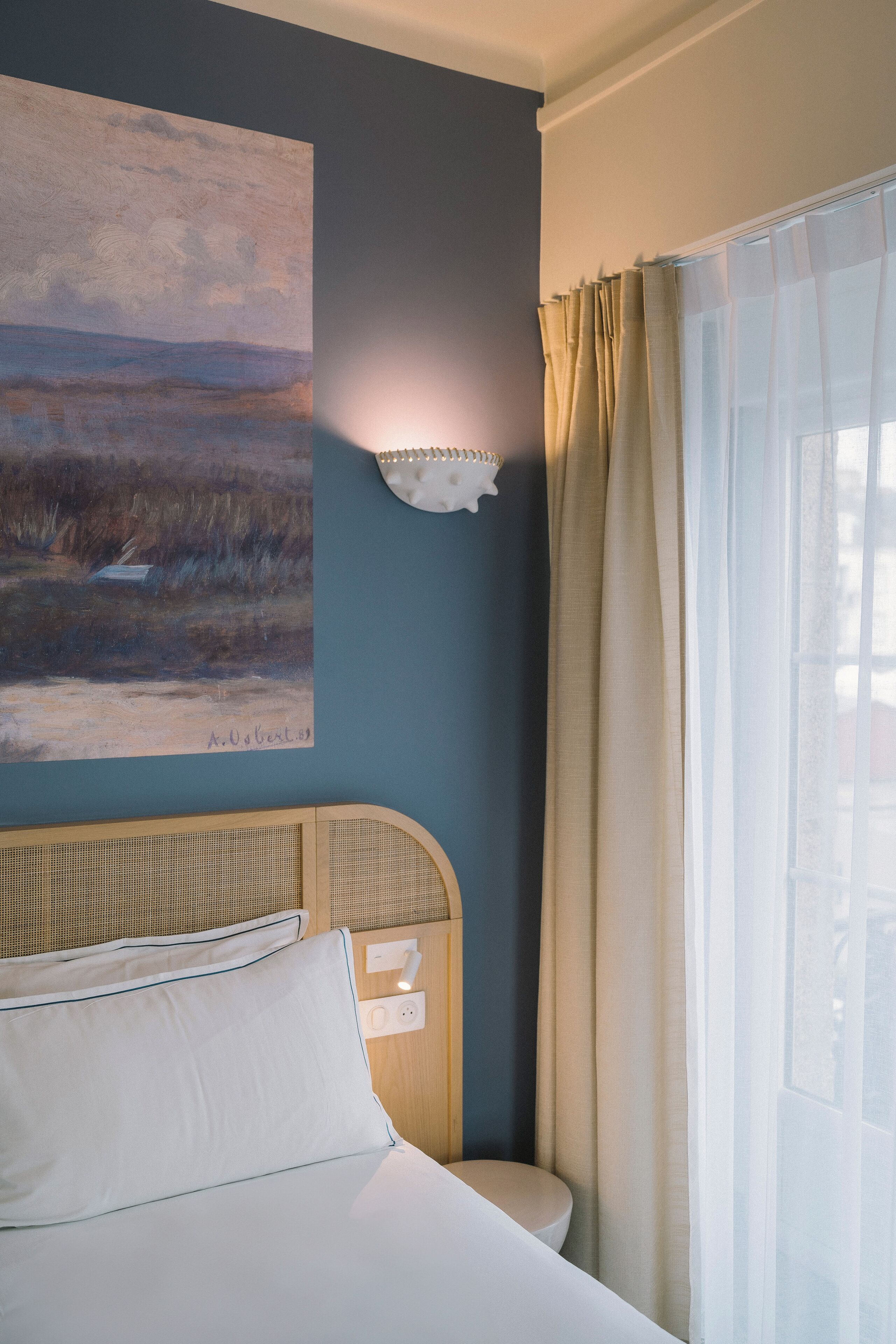 Ein Schlafzimmer mit einem Bett, einer Wandleuchte, einem Gemälde und einem Fenster mit Vorhängen.