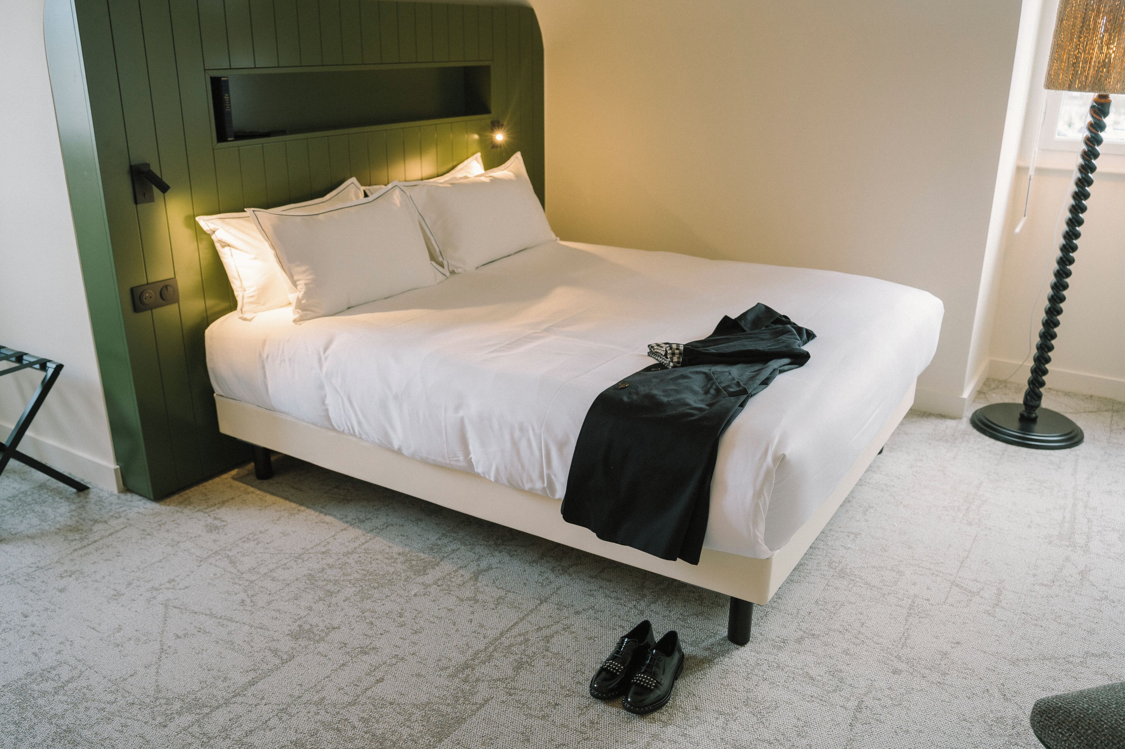 A neatly made bed with white linens and a black throw blanket, a pair of shoes on the floor, and a lamp with a twisted base beside it.