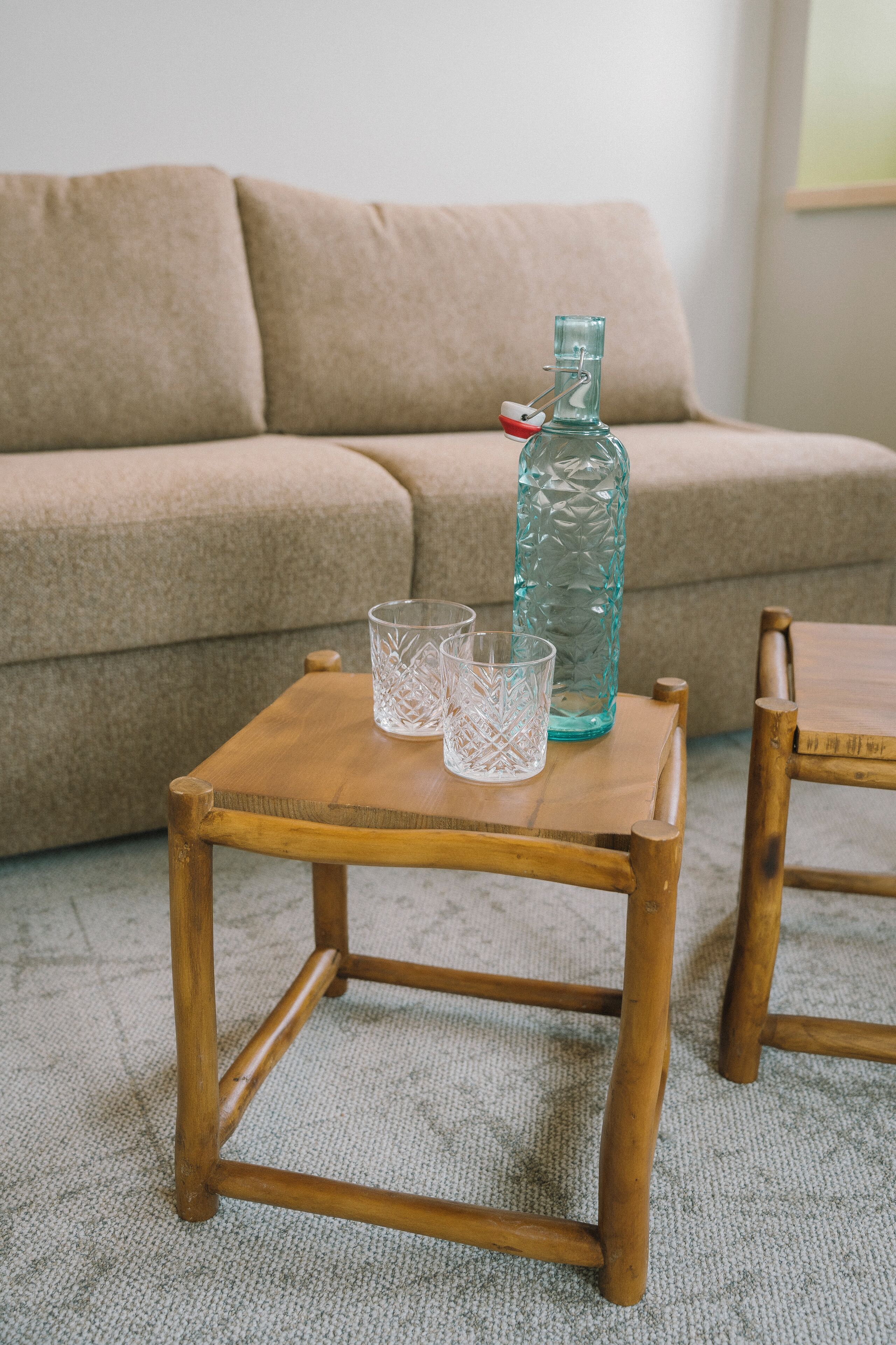 Een houten tafel met een glazen fles en twee glaasjes, geplaatst voor een beige bank.