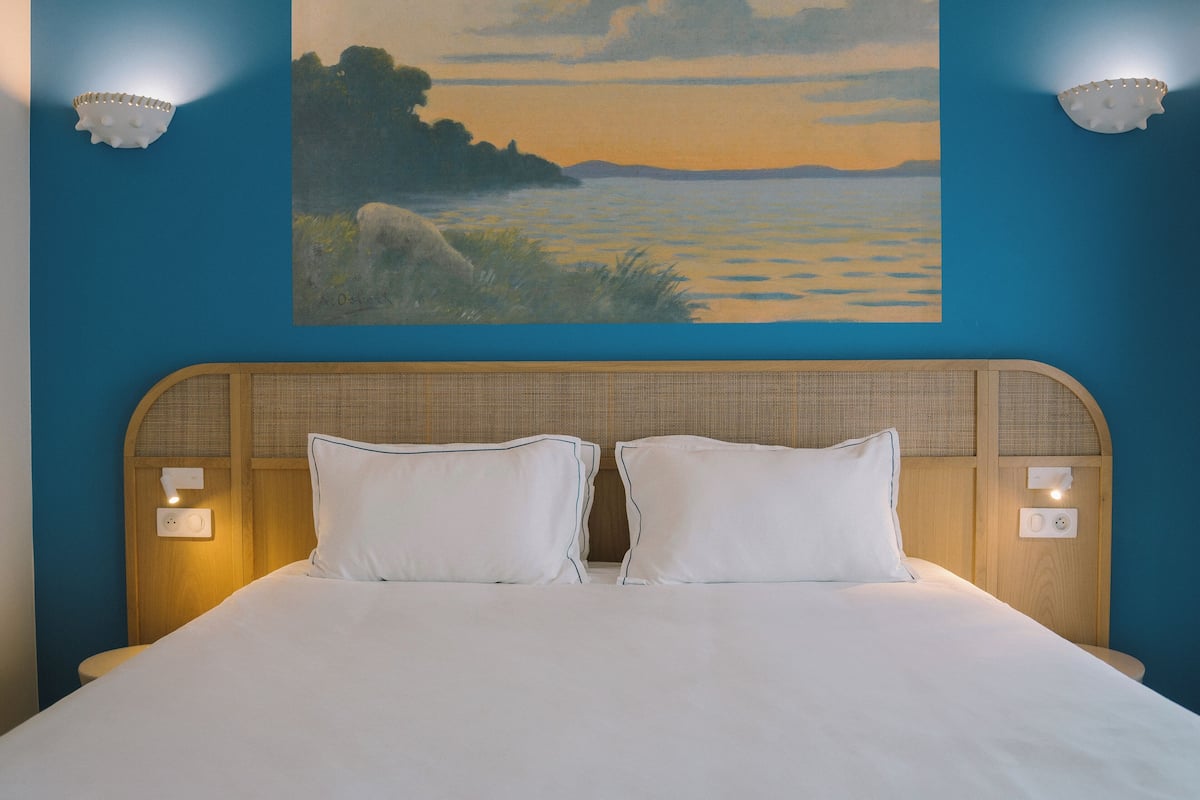 Cama doble con ropa de cama blanca, dos lámparas de noche y un cuadro de un paisaje en la pared.