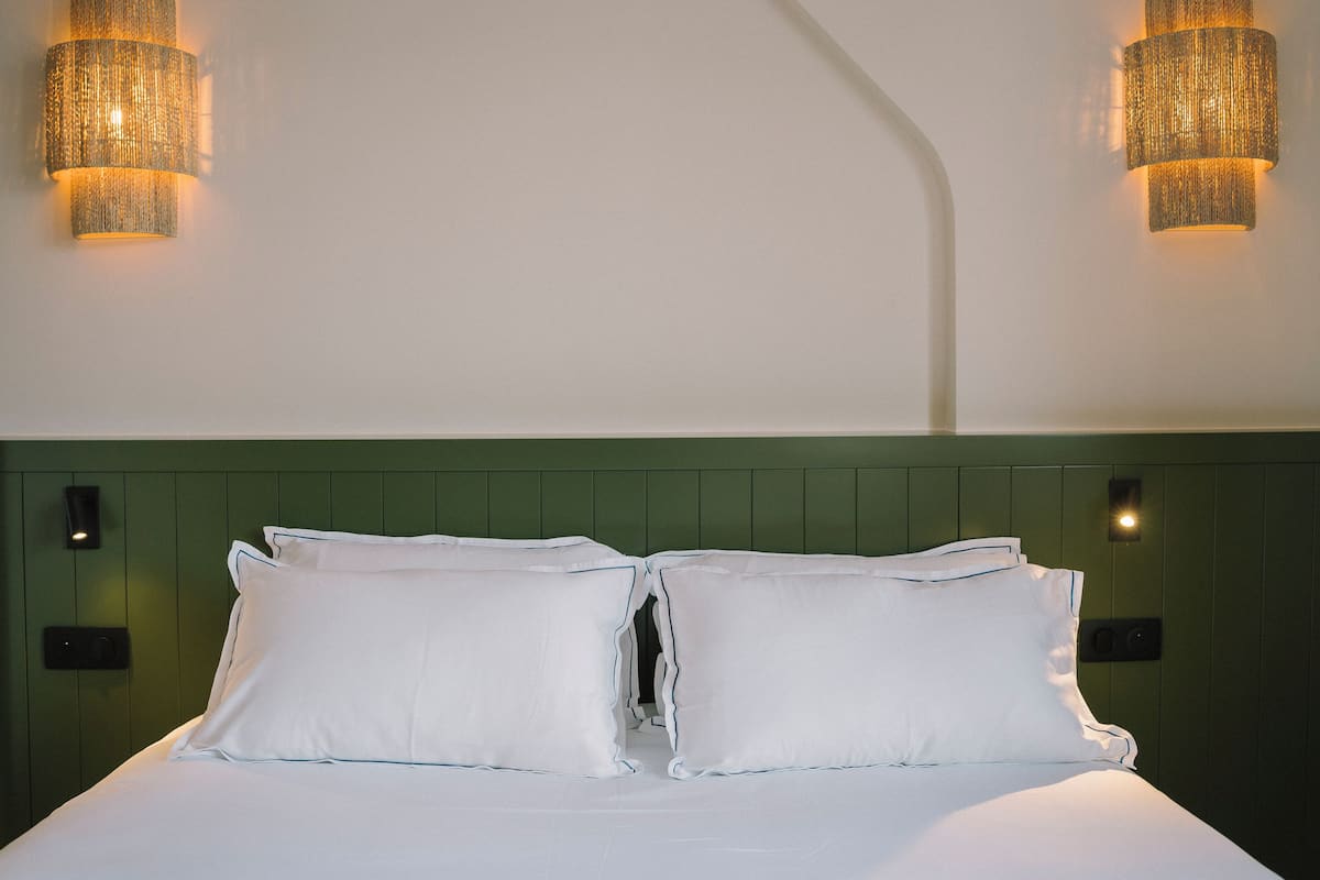 Una cama bien hecha con sábanas y almohadas blancas, un cabecero verde y dos lámparas fijadas a la pared.