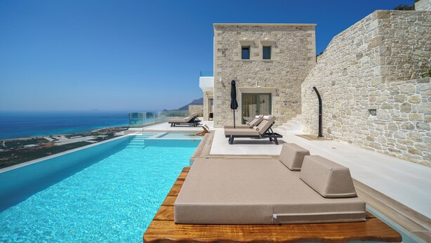 Un espace aménagé au bord de la piscine, avec des chaises longues et un bâtiment en pierre, offrant une vue sur une mer d’un bleu limpide.