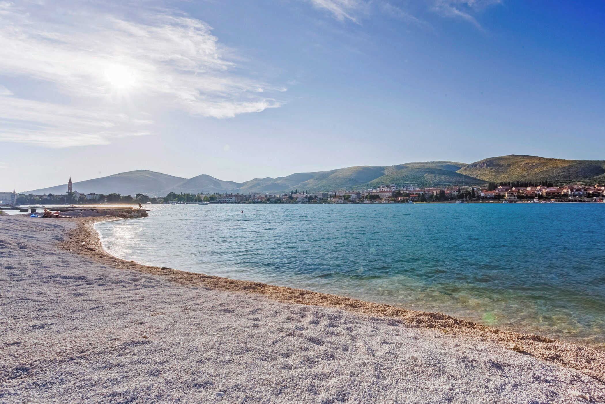 A beach with clear water, a pebble shore, and a distant town with mountains.