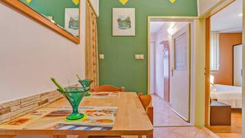 A dining area with a wooden table, chairs, and a green glass pitcher. There are framed pictures on the wall and a door leading to another room.