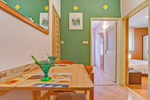 Ein Essbereich mit einem Holztisch, Stühlen und einer grünen Glas-Kanne. An der Wand befinden sich gerahmte Bilder und eine Tür, die in einen anderen Raum führt.