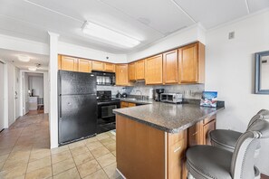 A kitchen with wooden cabinets, a black refrigerator, a granite countertop, and a coffee maker. - Marigot Beach 108 Oceanfront (Ocean City)