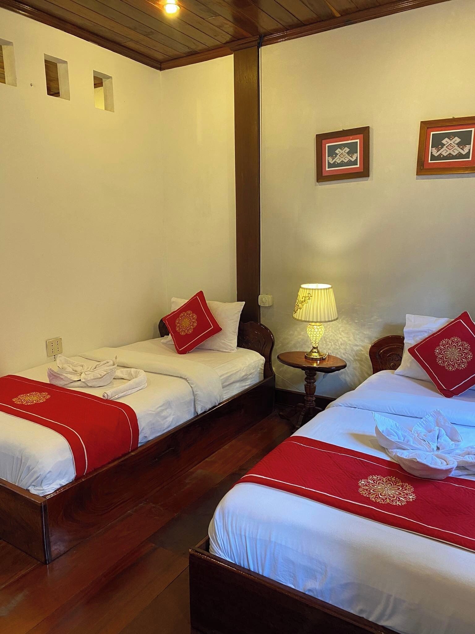 Two single beds with red headboards and white bedding, a wooden floor, a small table with a lamp, and a wall with two framed pictures.