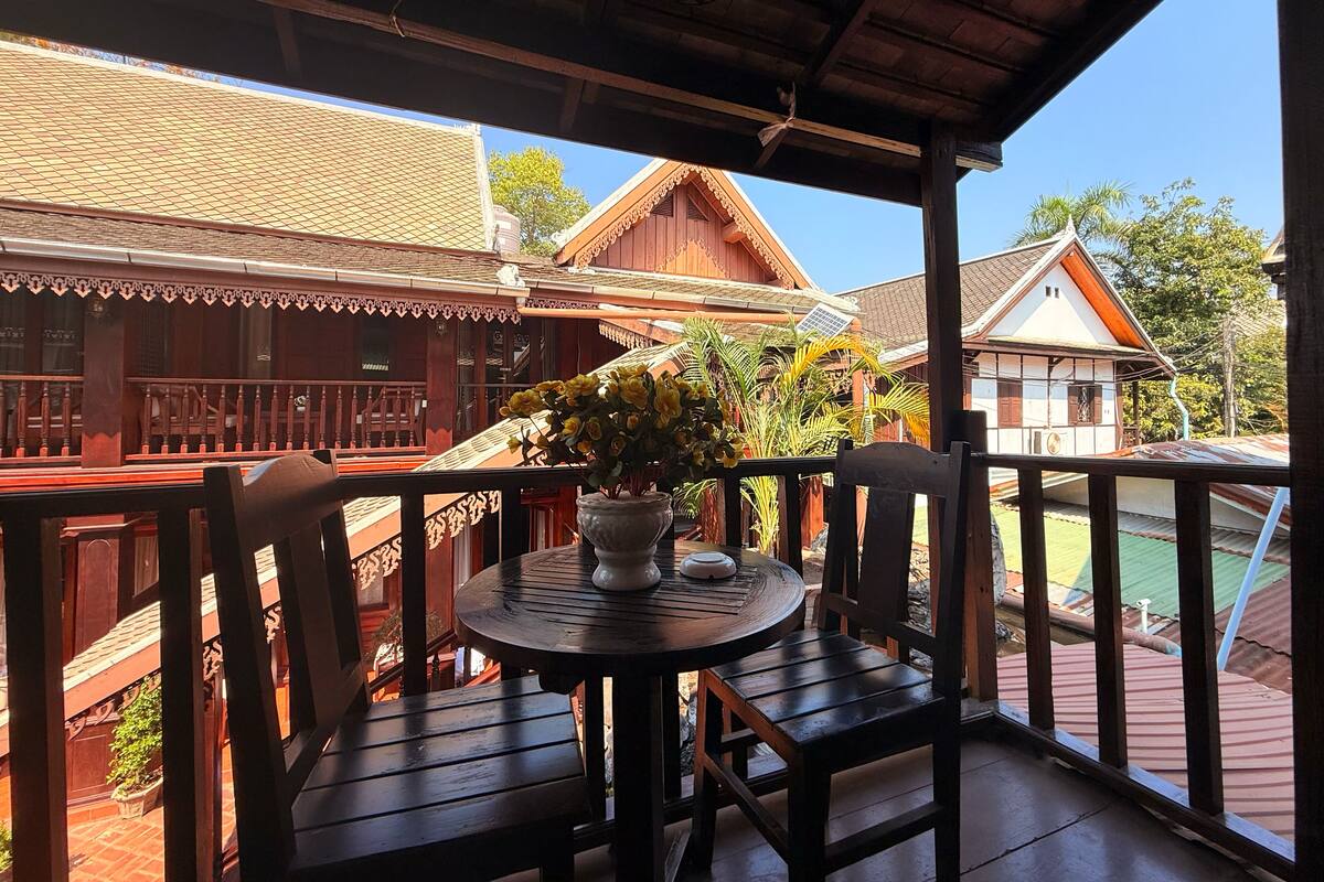 A wooden balcony with a round table and chairs, overlooking traditional buildings and a clear sky.