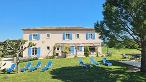 A two-story building with a tiled roof, blue shutters, and a garden with lawn chairs. - Maison d'hôtes La Ramade, 5 chambres et table d'hôtes en Provence (12 pers) (Tulette)