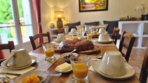 A breakfast setting with a wooden table, plates, glasses, a loaf of bread, and a painting on the wall. - Maison d'hôtes La Ramade, 5 chambres et table d'hôtes en Provence (12 pers) (Tulette)