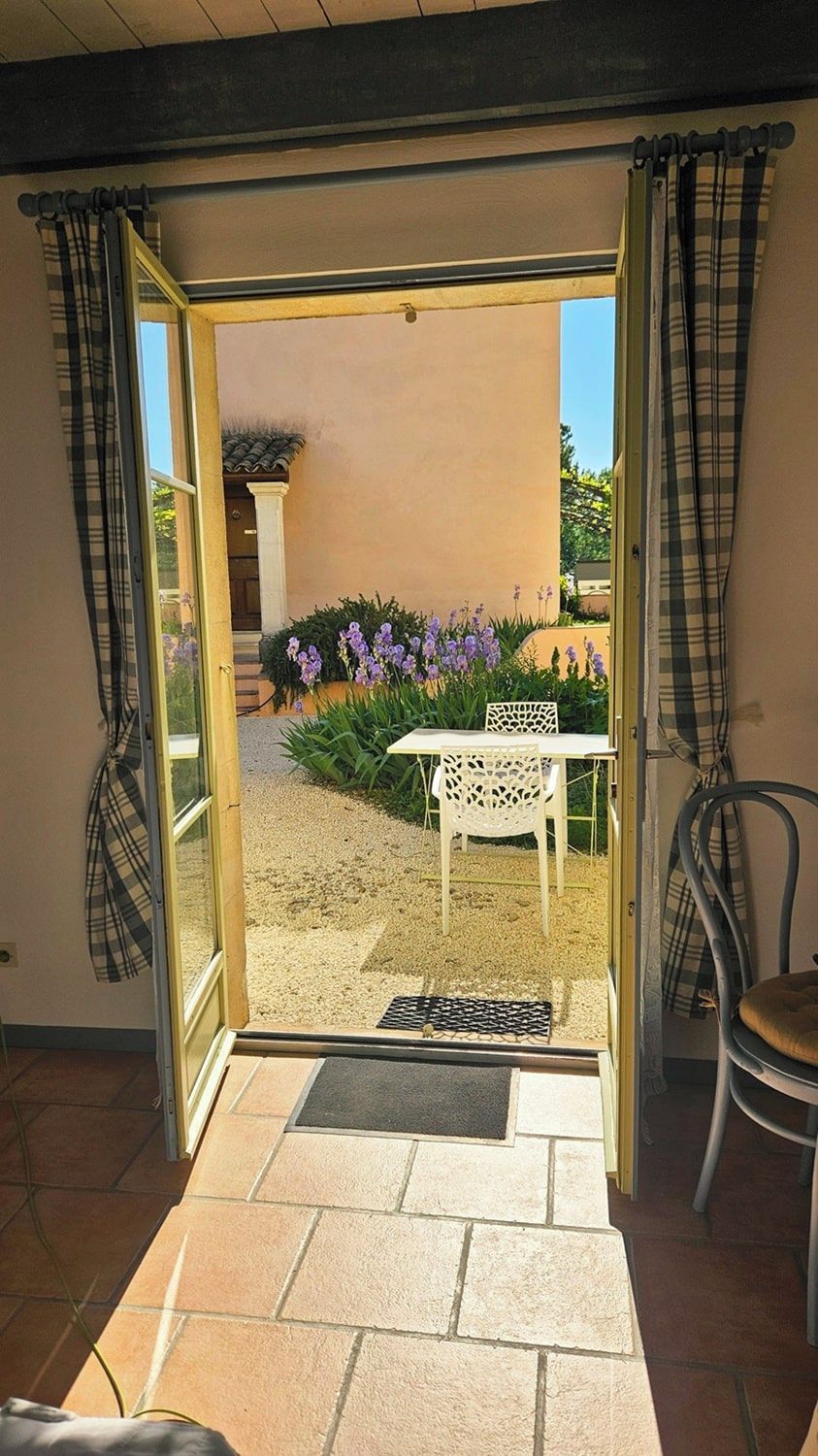 Una puerta corrediza de vidrio abierta que da a un patio con una mesa y sillas blancas, rodeado de flores moradas y un pequeño jardín.