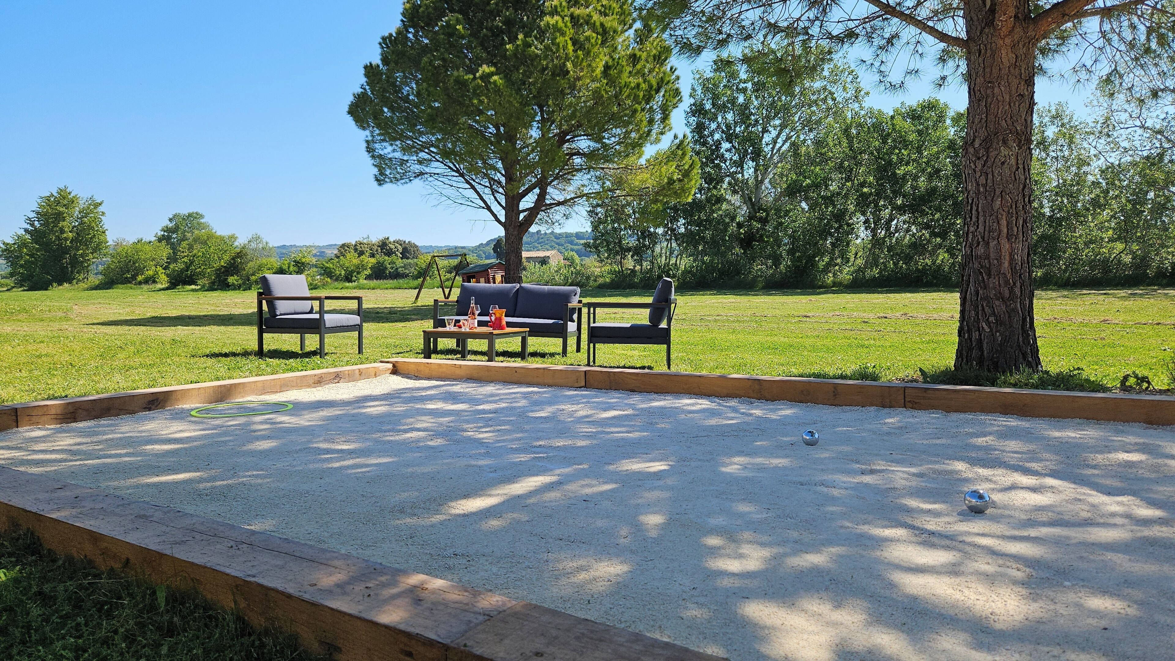 Un terrain de pétanque délimité par une bordure en bois, avec deux chaises, une table et une balançoire installée dans un champ herbeux.