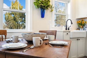 Una mesa de comedor con platos, tazones y tazas, un jarrón azul con hojas verdes, y una ventana con vista a árboles.