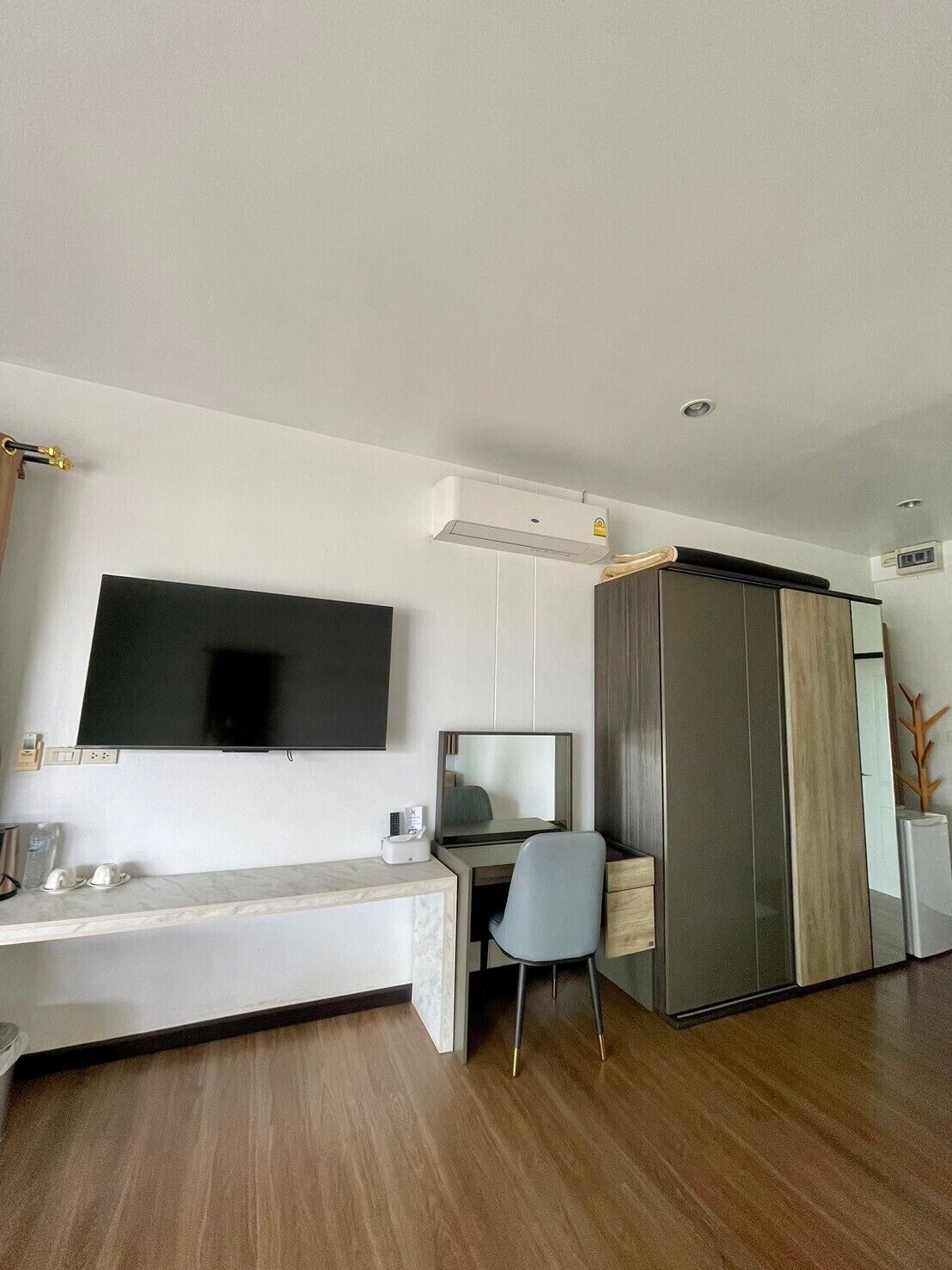 A modern hotel room with a flat-screen TV mounted on the wall, a desk with a chair, a wardrobe, and an air conditioning unit.