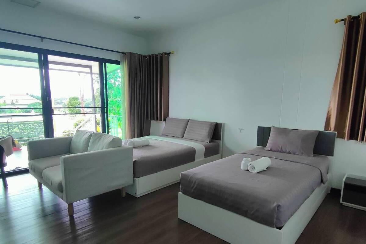 A modern hotel room with two single beds, a sofa, and a large window offering a view of the outdoors.
