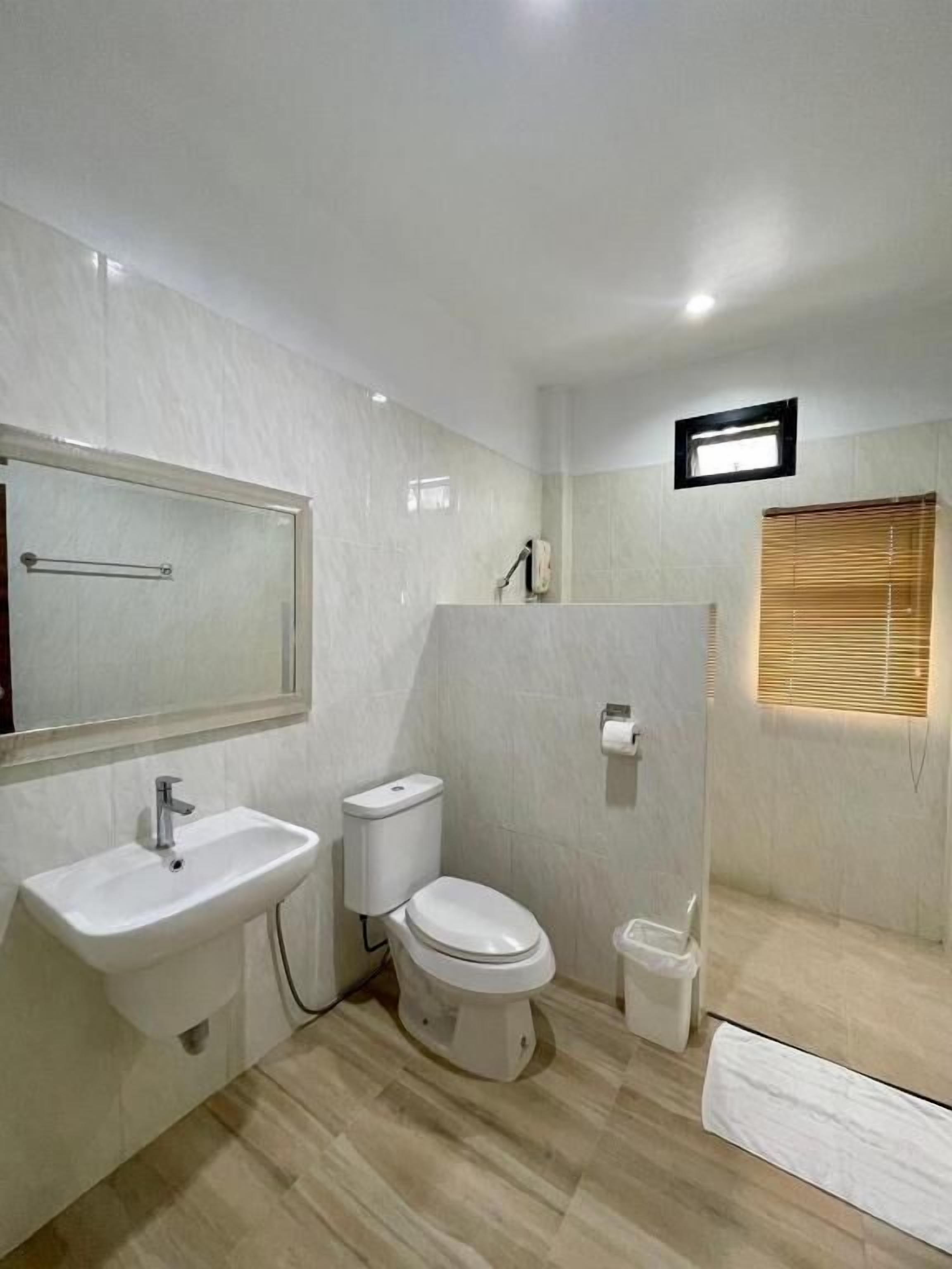 Une salle de bain moderne équipée d’un lavabo, de toilettes et d’un bidet. On y trouve un miroir, une petite fenêtre avec des stores et un luminaire au plafond.