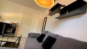 Ein modernes Wohnzimmer mit einem grauen Sofa, einem Glastisch, einer schwarzen Pendelleuchte und einem schwarzen Regal mit einer kleinen Pflanze.