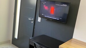 Ein Zimmer mit schwarzem Schreibtisch, einem an der Wand montierten Fernseher, einer Tür mit Lichtleiste und einem kleinen Holztisch.