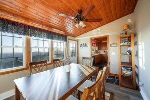 Un espace repas avec une longue table en bois, des chaises, un ventilateur de plafond et de grandes fenêtres offrant une vue sur les montagnes.