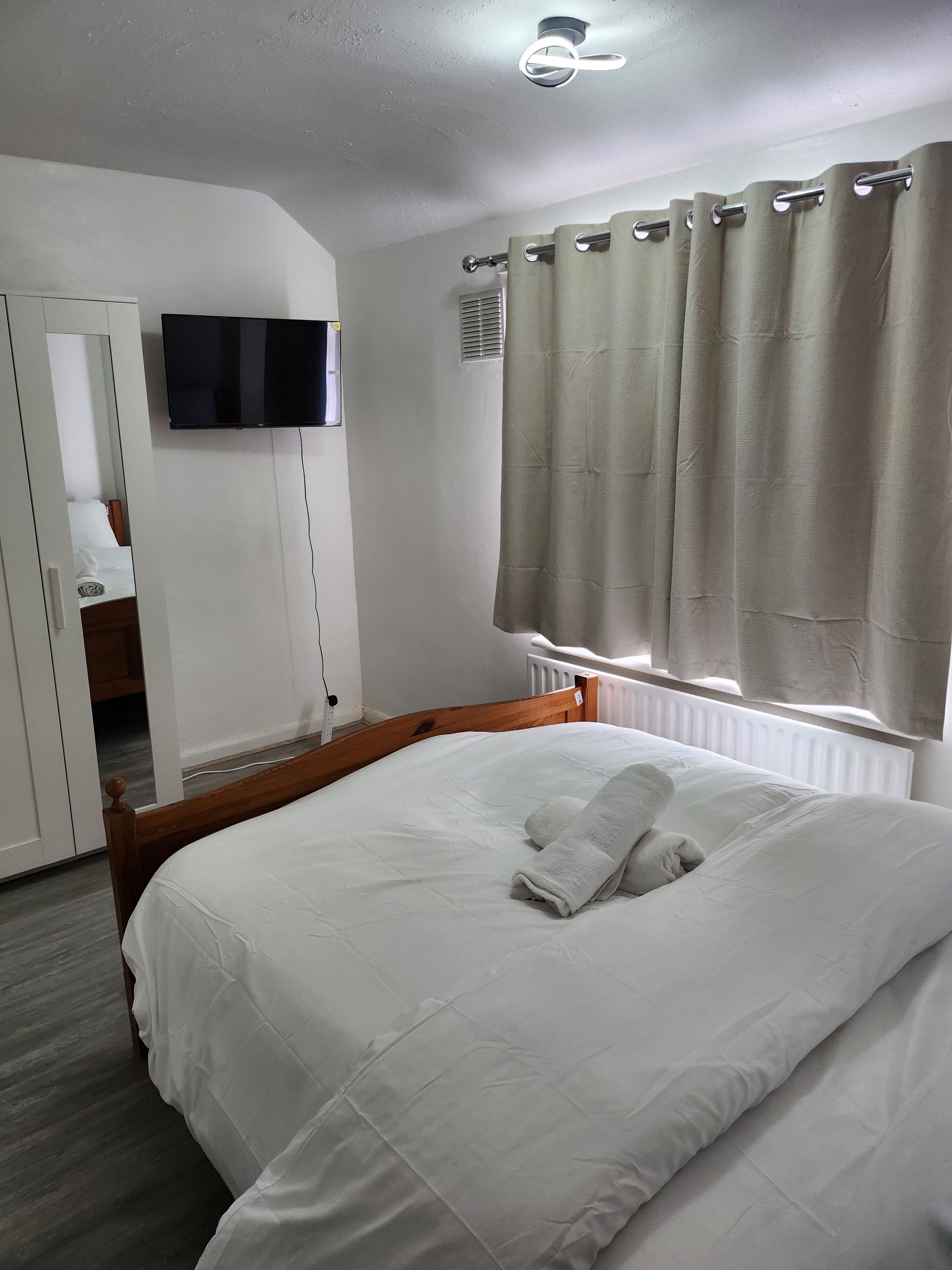 Un lit bien fait, avec du linge de lit blanc et une serviette enroulée, un téléviseur fixé au mur et une fenêtre ornée de rideaux beiges.
