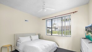 A bedroom with a bed, a ceiling fan, a window with bars, a small table, and a TV stand.