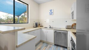 A modern kitchen with white cabinets, a dishwasher, a sink, and a window with a blue shade. A sign on the wall reads "meet me at the beach".
