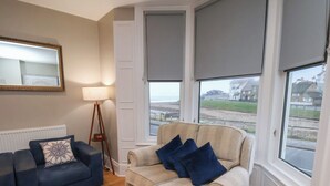 Ein Wohnzimmer mit einem beigefarbenen Sofa, einem blauen Sofa und einem Holztisch. Eine Lampe, eine Uhr und Blick durch die Fenster auf den Strand sind zu sehen.