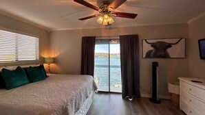 Un dormitorio con cama, ventilador de techo, ventana con cortinas, televisor y vista al agua.