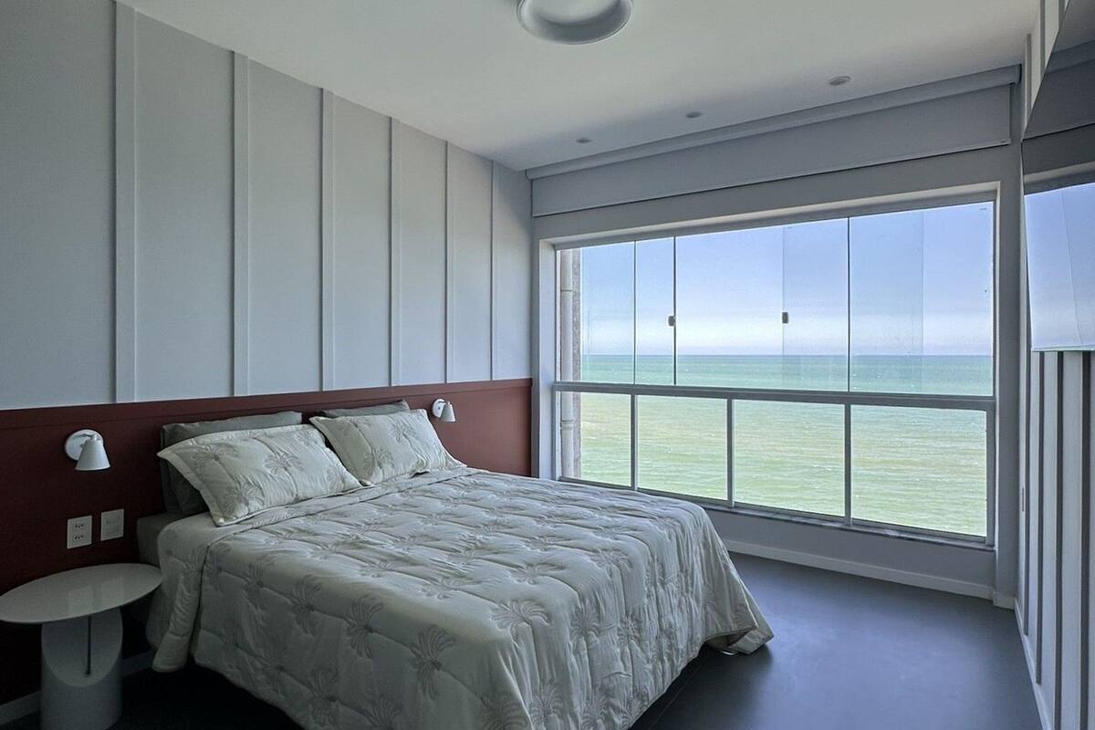 Un dormitorio moderno con una cama grande, una mesita de noche, una lámpara de pared y un ventanal con vistas al mar.