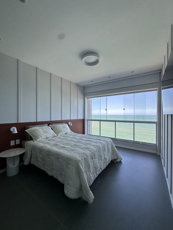 A modern bedroom with a large bed, a nightstand, a wall-mounted lamp, and a large window offering a view of the sea. - Guarapari Center Aparts (Guarapari)
