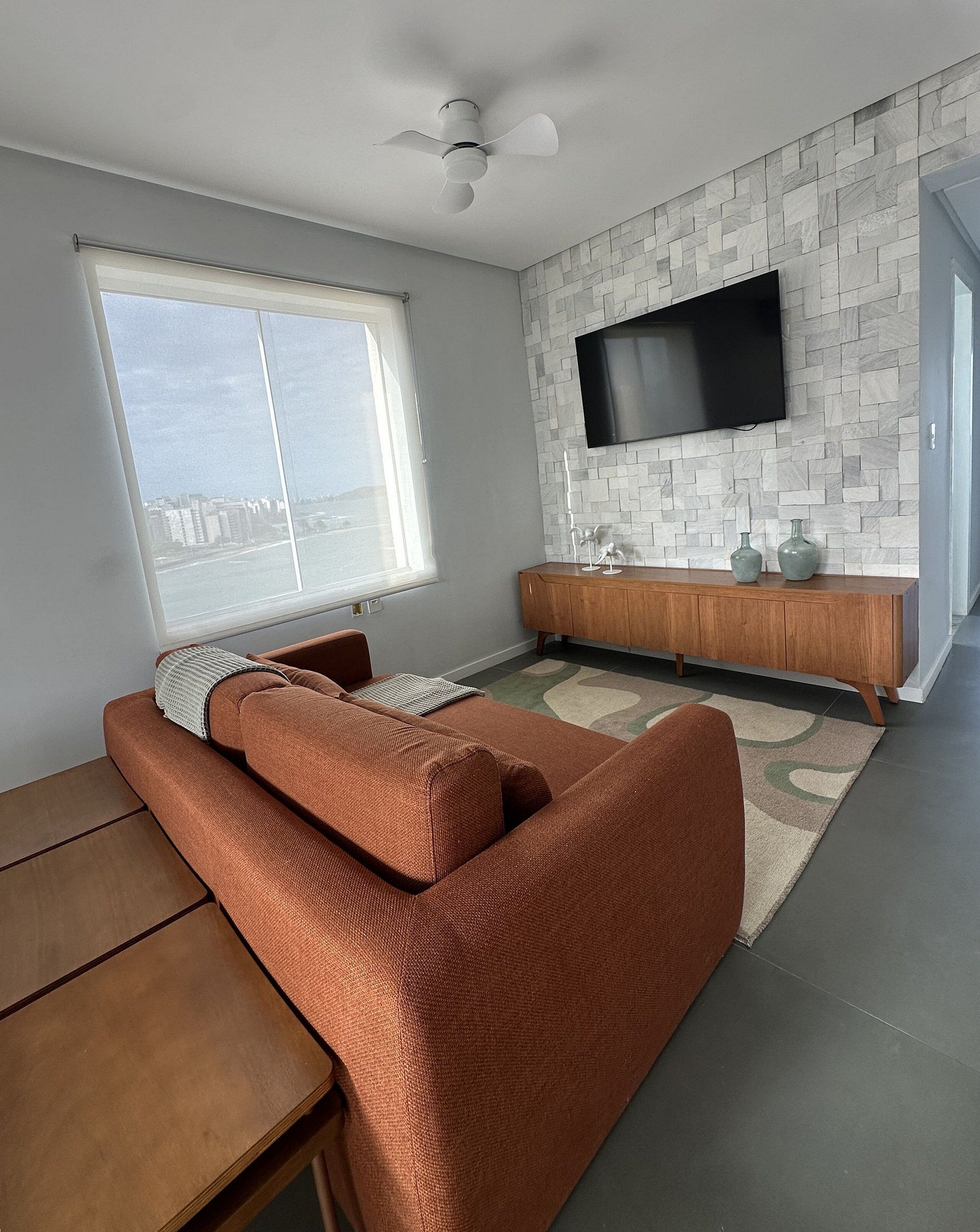 Una sala de estar moderna con un sofá, un televisor montado en una pared con diseño de piedra y vistas de la ciudad a través de un ventanal grande.