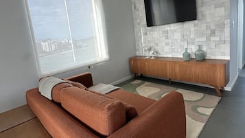 Una sala de estar moderna con un sofá, un televisor montado en una pared con diseño de piedra y vistas de la ciudad a través de un ventanal grande.