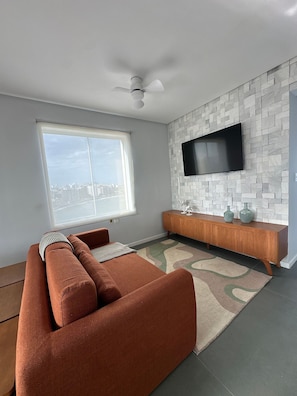A modern living room with a sectional sofa, a flat-screen TV mounted on a textured wall, a ceiling fan, and a large window offering a city view. - Guarapari Center Aparts (Guarapari)